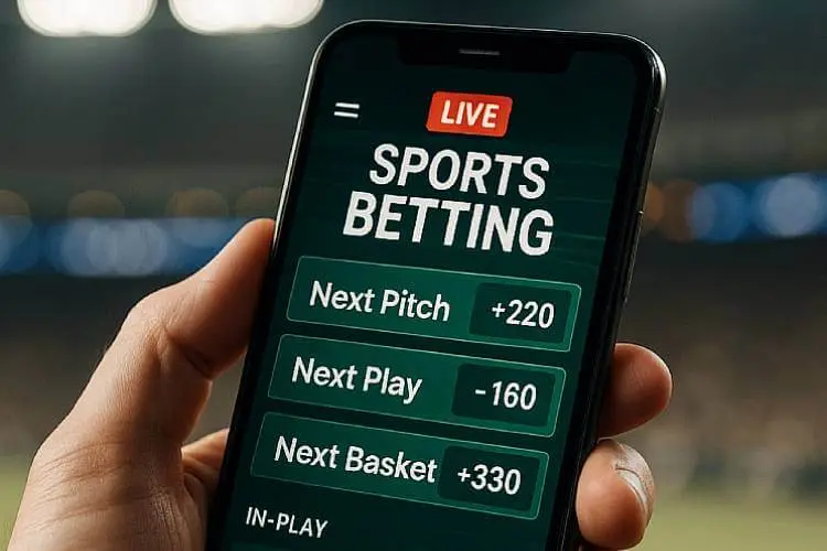 A smartphone held in a person’s hand displaying live microbetting options such as next pitch and next play, with a blurred stadium in the background.
